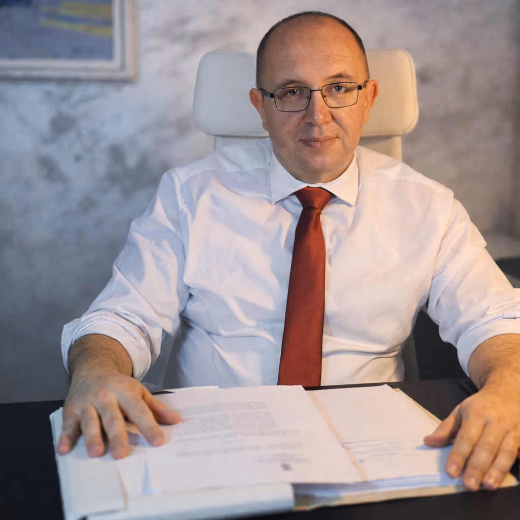 Lawyer wearing glasses and red tie sitting at desk with open documents, looking at camera.